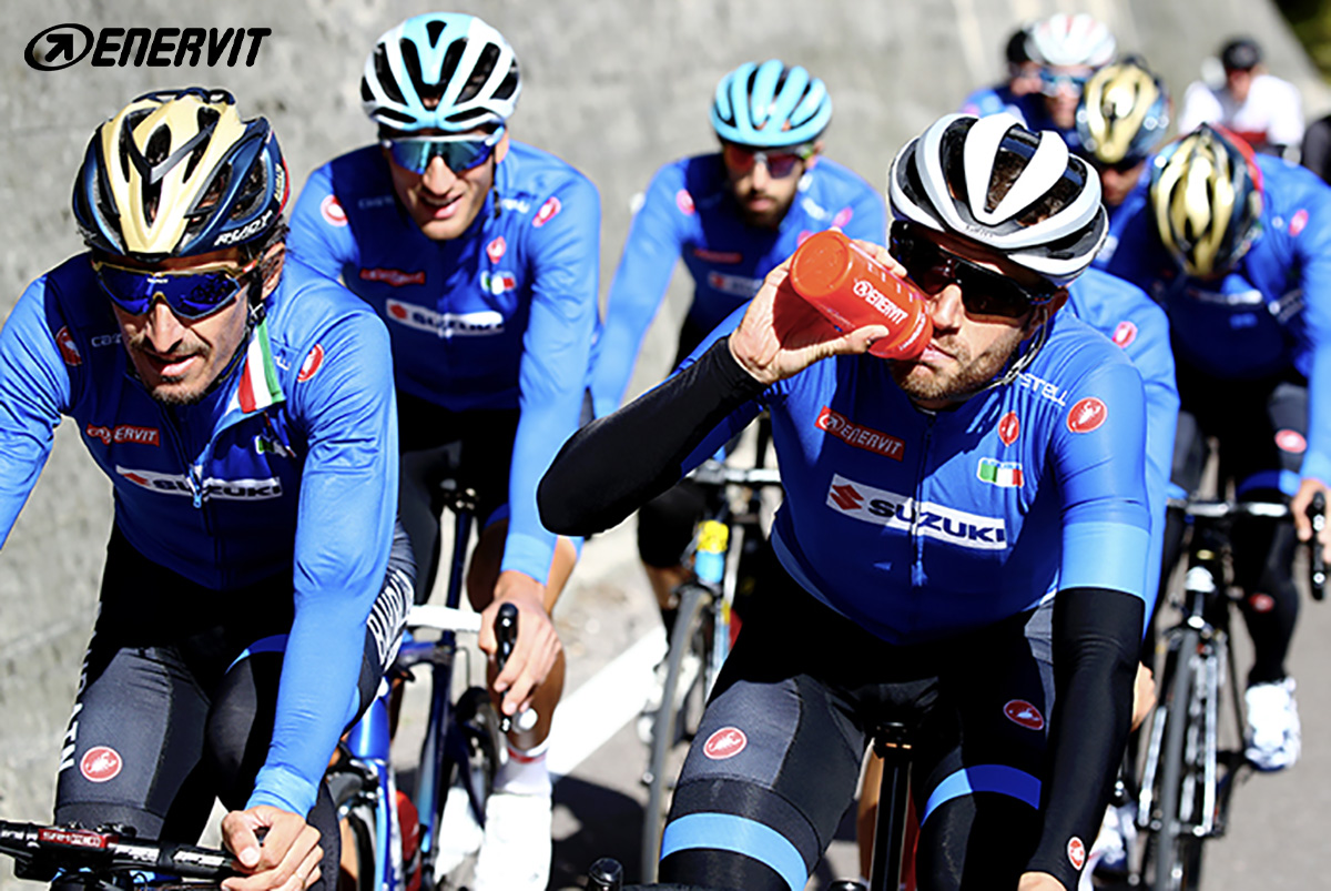 Gli atleti della nazionale italiana di ciclismo procedono in gruppo su una strada e si dissetano bevendo integratori Enervit